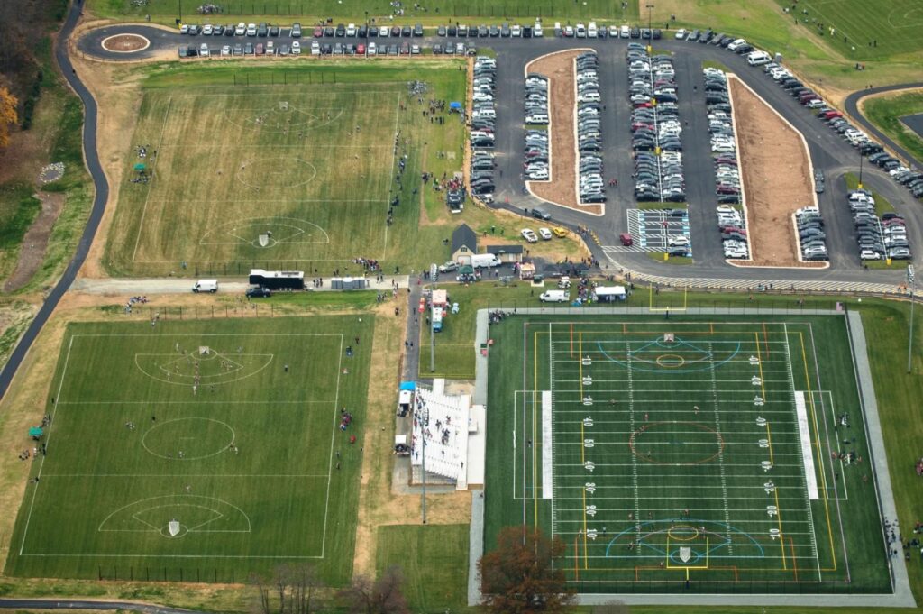 Aerial view of a large sports complex with multiple fields including synthetic turf football field, parking area, and active games in progress
